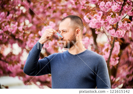 Man allergic suffering from seasonal allergy at spring. Bearded man with clothespin clipped to his nose - symbolic gesture of his inability to breathe due to nasal congestion near blooming tree. 121615142