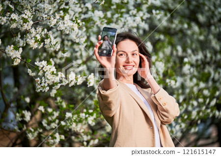 Happy woman taking selfie with smartphone among spring blossoms with soft focus on the background. 121615147