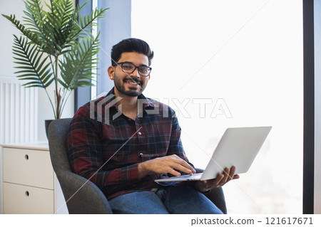 Smiling young Indian man sitting in office chair holding laptop. Casual work environment with natural light. Comfortable and relaxed atmosphere. Modern workspace with greenery. Smiling young Indian man sitting in office chair holding laptop. Casual work environment with natural light. Comfortable and relaxed atmosphere. Modern workspace with greenery. 121617671