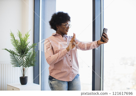 Young African American businesswoman taking selfie near large window. She wears glasses, casual outfit. Bright office setting conveys modern work environment, confidence, connection. Young African American businesswoman taking selfie near large window. She wears glasses, casual outfit. Bright office setting conveys modern work environment, confidence, connection. 121617696