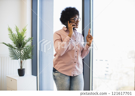 African American woman in casual attire engaged in phone conversation near window in office setting, conveying confidence and professionalism. 121617701