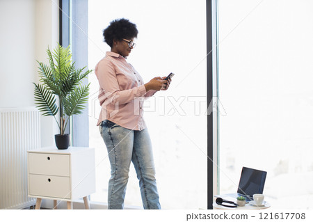 African American businesswoman in casual attire uses smartphone near window in bright office. Young female stands confidently with natural light creating warm atmosphere. 121617708