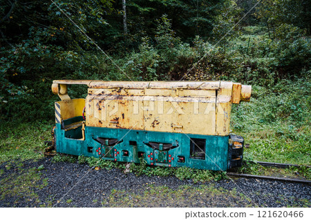 An abandoned mining locomotive sits peacefully among overgrown vegetation, showcasing its weathered body and colorful paint. This relic of industry contrasts with the surrounding forest. An abandoned mining locomotive sits peacefully among overgrown vegetation, showcasing its weathered body and colorful paint. This relic of industry contrasts with the surrounding forest. 121620466