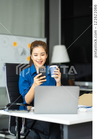 Asian businesswoman sits in a office working and enjoys a coffee. Asian businesswoman sits in a office working and enjoys a coffee. 121621066