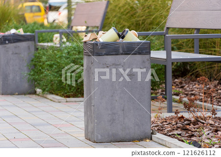 Rubbish piles up in a concrete waste container near park benches, showcasing a common sight of litter accumulation. The green surroundings underscore the need for better environmental practices 121626112