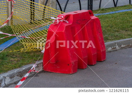 A vivid red plastic barrier stands prominently on the roadside, cordoned off with caution tape as workers prepare for upcoming construction. Sunlight illuminates the area, enhancing visibility 121626115