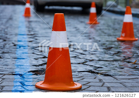 Colorful traffic cones provide clear direction on a wet and glossy street. Rain droplets enhance the environment, reflecting colors and creating patterns on cobblestones 121626127