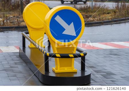A bright yellow directional sign with a blue circular background and white arrow points left in a rainy urban setting. The sign is placed on a pedestrian crossing area made of black paving stones A bright yellow directional sign with a blue circular background and white arrow points left in a rainy urban setting. The sign is placed on a pedestrian crossing area made of black paving stones 121626140
