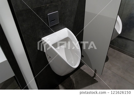 A modern urinal is mounted on a dark tiled wall in a well-lit public restroom. The minimalistic design emphasizes clean lines, creating a contemporary feel in the space 121626150