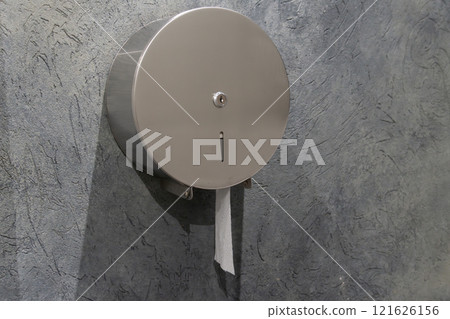 A round stainless steel toilet paper dispenser is securely mounted on a textured gray wall inside a public restroom. The dispenser has a small opening for easy paper access 121626156