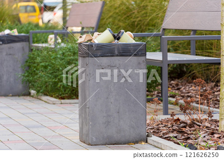 A concrete trash bin stands filled with waste bags, surrounded by greenery and benches in a bustling urban park. This spot reflects both convenience and environmental responsibility 121626250