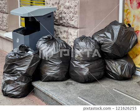 Several large black trash bags piled up beside a recycling bin on a city sidewalk. The setting emphasizes urban waste management and environmental conservation efforts in action Several large black trash bags piled up beside a recycling bin on a city sidewalk. The setting emphasizes urban waste management and environmental conservation efforts in action 121626255