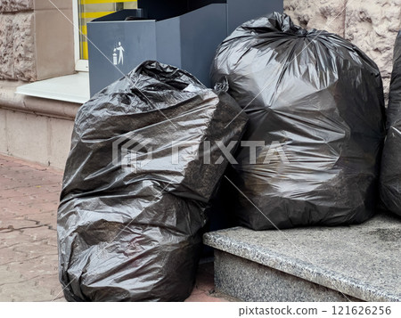 Black garbage bags lean against a clean, modern trash container on a city sidewalk. The sunlight highlights the need for environmental awareness and responsible waste management in urban areas Black garbage bags lean against a clean, modern trash container on a city sidewalk. The sunlight highlights the need for environmental awareness and responsible waste management in urban areas 121626256