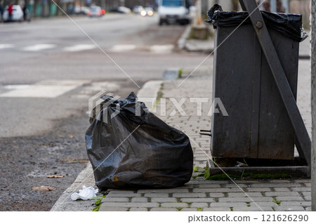 Two black trash bags rest beside a metallic garbage container along a city sidewalk, emphasizing the ongoing issues with urban waste disposal and environmental conservation efforts Two black trash bags rest beside a metallic garbage container along a city sidewalk, emphasizing the ongoing issues with urban waste disposal and environmental conservation efforts 121626290