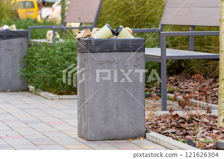 A concrete garbage container is overflowing with trash bags and recyclables in a well-maintained urban park. Nearby benches and greenery enhance the outdoor atmosphere, highlighting waste management 121626304