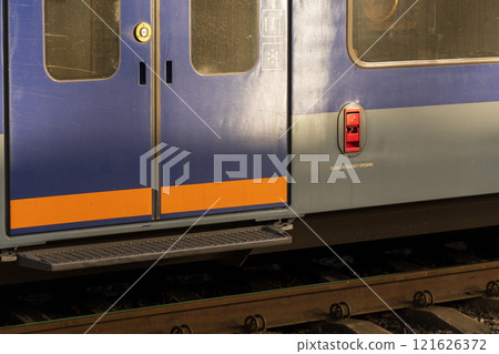 The sleek design of modern electric train doors stands out at a railway station, illuminated by warm light from the setting sun. Trains await passengers on the platform The sleek design of modern electric train doors stands out at a railway station, illuminated by warm light from the setting sun. Trains await passengers on the platform 121626372