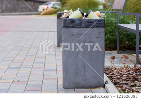 A concrete garbage container is filled with trash bags on a busy city sidewalk. Nearby plants and benches are visible, highlighting urban waste management challenges A concrete garbage container is filled with trash bags on a busy city sidewalk. Nearby plants and benches are visible, highlighting urban waste management challenges 121626385