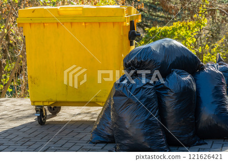 Black garbage bags are arranged next to a yellow waste container in a bright outdoor setting. The scene highlights waste disposal practices in an urban environment Black garbage bags are arranged next to a yellow waste container in a bright outdoor setting. The scene highlights waste disposal practices in an urban environment 121626421