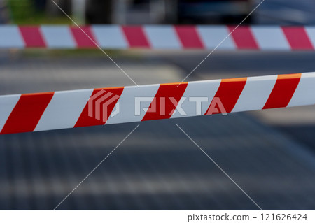 Caution tape with bold red and white stripes stretches across a pathway, clearly marking a restricted area in a busy city setting. The sun casts shadows on the pavement 121626424