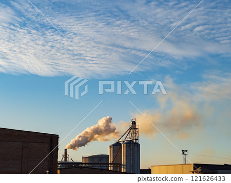 Thick smoke rises from multiple smokestacks in an industrial area as the sun sets, casting a warm glow on the surrounding buildings and machinery Thick smoke rises from multiple smokestacks in an industrial area as the sun sets, casting a warm glow on the surrounding buildings and machinery 121626433