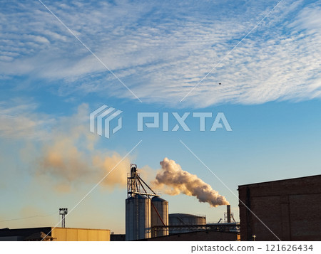Billowing smoke emerges from smokestacks at an industrial facility during sunset, casting a cloudy backdrop over the manufacturing buildings and machinery 121626434