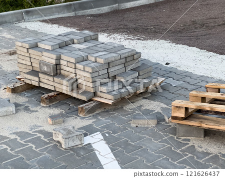 Sturdy concrete pavers are stacked on wooden pallets, surrounded by loose sand and gravel at an urban construction site near a paved street, ready for installation Sturdy concrete pavers are stacked on wooden pallets, surrounded by loose sand and gravel at an urban construction site near a paved street, ready for installation 121626437