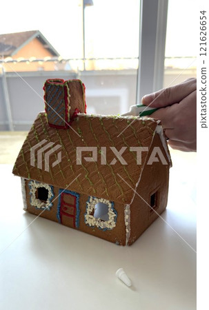 Hand Decorating Festive Gingerbread House with Colorful Icing in Bright Setting 121626654