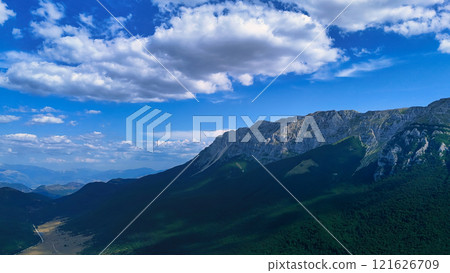 Majestic mountain range casting shadows over green valley under blue sky Majestic mountain range casting shadows over green valley under blue sky 121626709