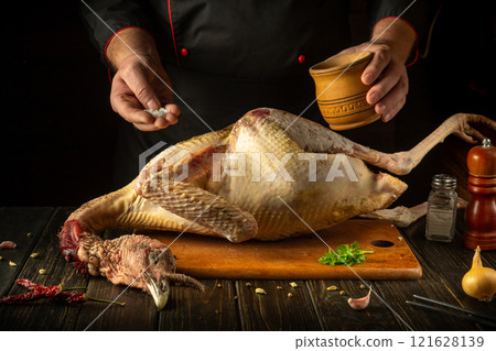 A skilled chef expertly sprinkles salt on a large raw turkey on a wooden cutting board. The warm atmosphere of a country kitchen adds to the culinary creativity A skilled chef expertly sprinkles salt on a large raw turkey on a wooden cutting board. The warm atmosphere of a country kitchen adds to the culinary creativity 121628139