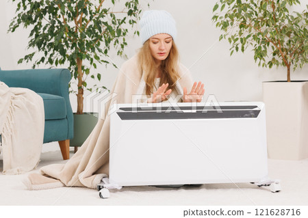 Young woman in socks with warm plaid near electric heater at home. Concept of heating season. Young woman in socks with warm plaid near electric heater at home. Concept of heating season. 121628716