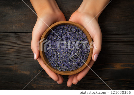 Hands gently holding a wooden bowl filled with dried lavender flowers on a rustic wooden surface 121630396