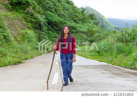 Portrait of a tired young Filipina woman walking along a road among mountains with a wooden stick 121631802