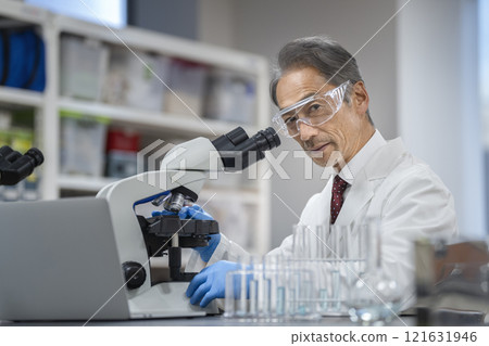 Senior man in white coat working with microscope, looking at camera, portrait Senior man in white coat working with microscope, looking at camera, portrait 121631946