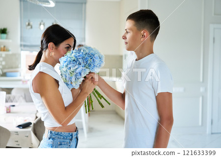 A boy surprises a girl with a bouquet of blue flowers in a modern kitchen during daylight A boy surprises a girl with a bouquet of blue flowers in a modern kitchen during daylight 121633399