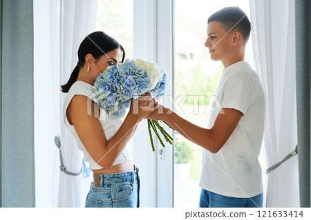 A young man surprises a woman with a bouquet of blue and white flowers indoors 121633414