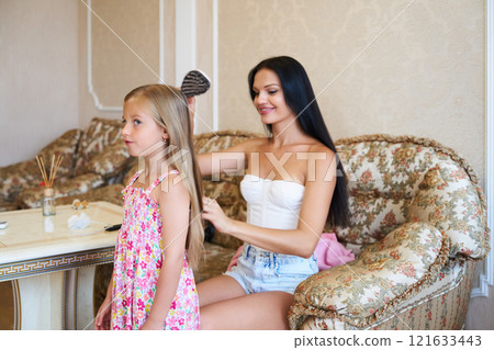 Mother Gently Brushes Her Daughter's Long Hair While Relaxing at Home in the Afternoon Light 121633443