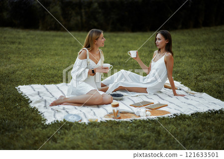 Friends enjoying tea on a picnic blanket in a lush green park setting 121633501
