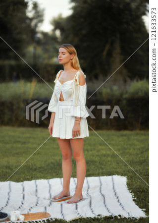 Model poses gracefully in a white dress on a blanket in a grassy area Model poses gracefully in a white dress on a blanket in a grassy area 121633573