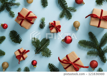 Festive background with small gift boxes wrapped in kraft paper, with bows of red velvet ribbon, bright green pine sprigs and red and gold Christmas ornaments. Blue backdrop. 121636980