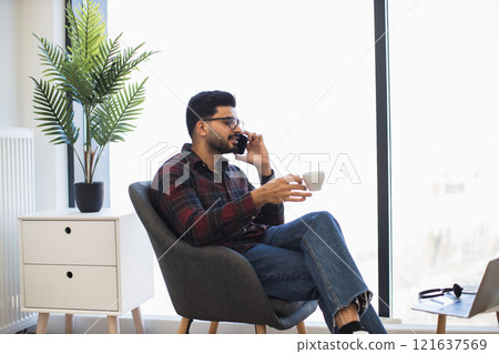 Indian man relaxing at home with coffee and phone, wearing casual clothes. Bright interior with modern decor, window light, and plant. Concept of leisure, communication, comfort, tranquility. Indian man relaxing at home with coffee and phone, wearing casual clothes. Bright interior with modern decor, window light, and plant. Concept of leisure, communication, comfort, tranquility. 121637569