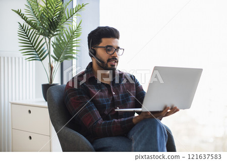 Young Indian man working remotely with laptop and headset, sitting comfortably. Engaged in online communication, focused on professional tasks. 121637583