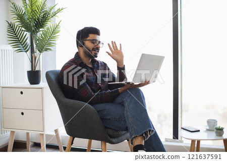 Indian young man sitting in chair having video call on laptop. Man wearing headset and plaid shirt, appears engaged and happy. Working from home concept. Indian young man sitting in chair having video call on laptop. Man wearing headset and plaid shirt, appears engaged and happy. Working from home concept. 121637591