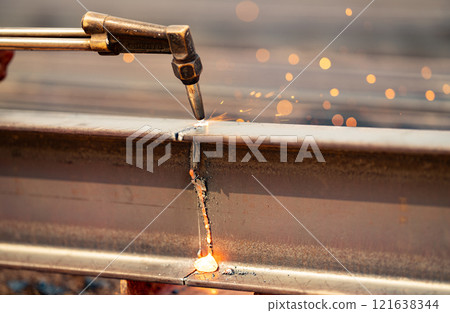 Welder using gas torch to cut metal beam, with sparks flying. Skilled craftsmanship and risks involved in industrial metal fabrication. Precision and danger of process. Cutting steel beam with a torch 121638344
