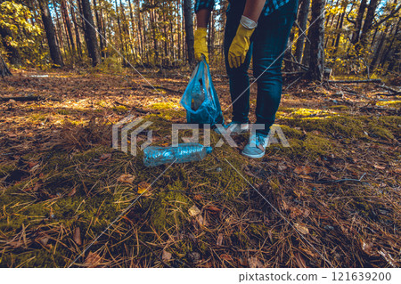 A gloved hand picks up a plastic bottle from the ground and places it in a trash bag. A volunteer man removes trash scattered in the forest. Eco, saving nature, eco activism. 121639200
