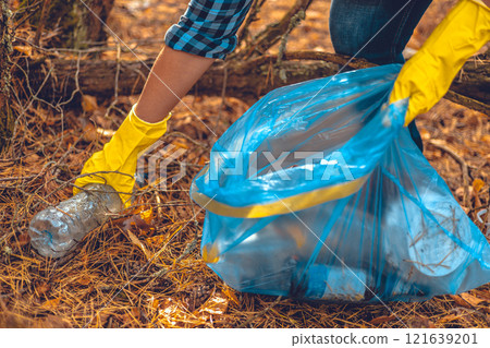 A woman conservationist collects garbage in the forest. A woman with a blue garbage bag in her hands collects plastic waste in the forest. A woman saves the forest from being polluted by garbage. A woman conservationist collects garbage in the forest. A woman with a blue garbage bag in her hands collects plastic waste in the forest. A woman saves the forest from being polluted by garbage. 121639201
