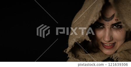 A grinning witch peeks out from under a hood on Halloween night. Copy space. Banner, flyer. A beautiful witch with a hood thrown over her head smirks while looking at a vammer on a black background. A grinning witch peeks out from under a hood on Halloween night. Copy space. Banner, flyer. A beautiful witch with a hood thrown over her head smirks while looking at a vammer on a black background. 121639306