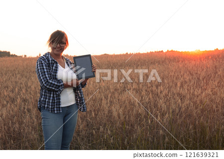 Against the backdrop of a beautiful sunset in a wheat field a female farmer smiles and looks at the camera with a tablet Copy space. A woman shows a tablet on the background of a field of ripe wheat. 121639321