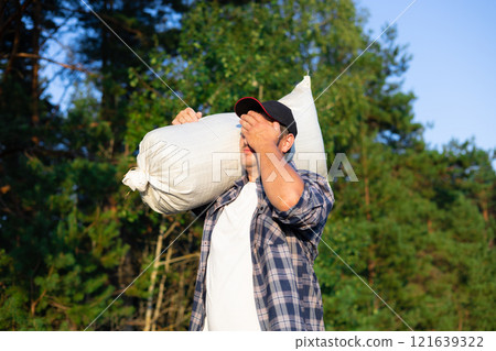 Wiping the sweat from his forehead with his hand, the farmer carries the harvested crop in a bag on his shoulder. Against the backdrop of a blue sky and a farmer man carries the harvest. Wiping the sweat from his forehead with his hand, the farmer carries the harvested crop in a bag on his shoulder. Against the backdrop of a blue sky and a farmer man carries the harvest. 121639322