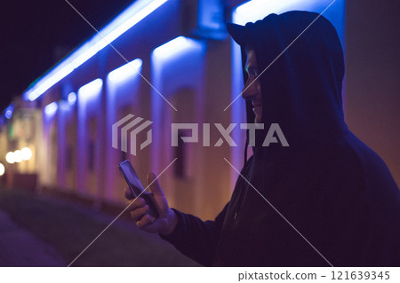 Night city life. A man with a phone in his hand stands at night on a city street illuminated by neon lights. Cyber punk. A man writes a message on his phone on a city street at night. Night city life. A man with a phone in his hand stands at night on a city street illuminated by neon lights. Cyber punk. A man writes a message on his phone on a city street at night. 121639345