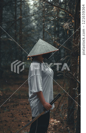 A woman wearing a straw hat and mask, depicting a samurai, stands in a forest, holding a katana at her side. The image evokes a sense of mystery and adventure. Woman with a katana. A woman wearing a straw hat and mask, depicting a samurai, stands in a forest, holding a katana at her side. The image evokes a sense of mystery and adventure. Woman with a katana. 121639364